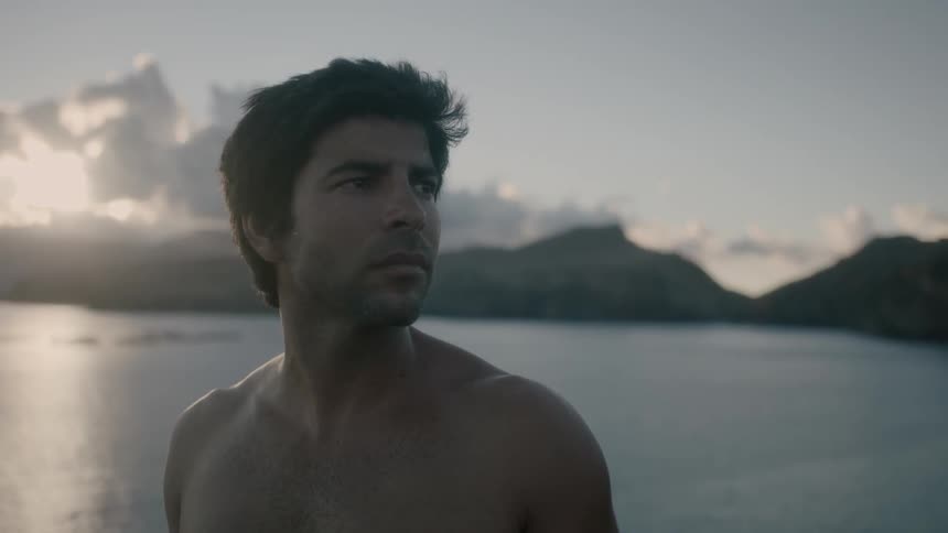 A shirtless male stares off to the side as he stands above a body of water and hills
