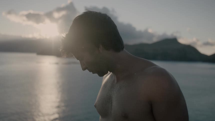 Shirtless man looking down with water and land behind him