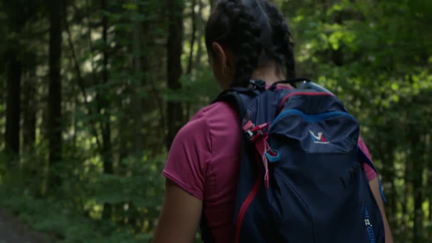 A woman with pig tails in her hair and wearing a backpack is walking through the woods as the sun shines through the trees