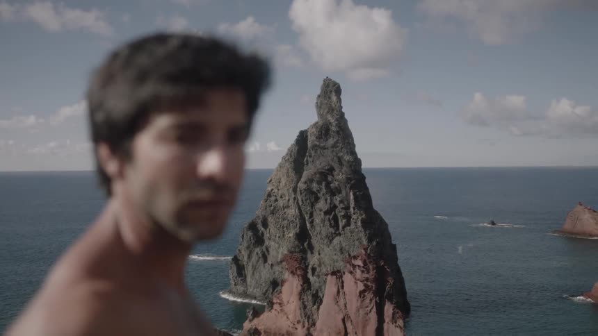 A shirtless man stands near the ocean in the daytime