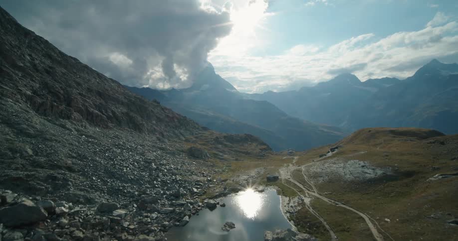 Trails move through valley around reflective lake near mountains leading toward foggy mountain range under a cloudy sky
