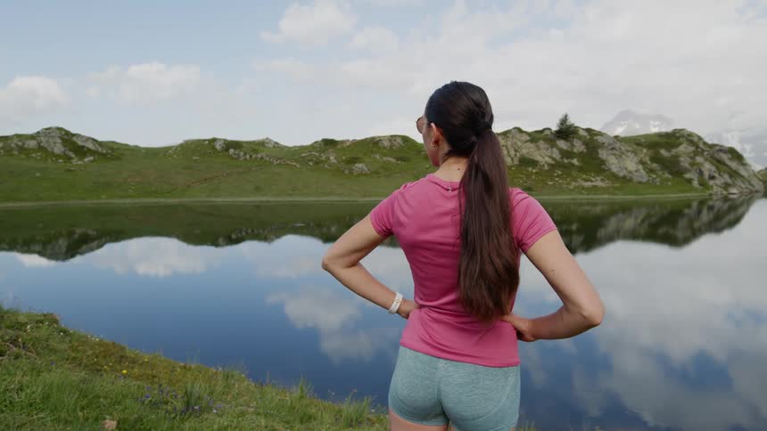 Woman stands across from mountains on shore of lake reflecting clouds