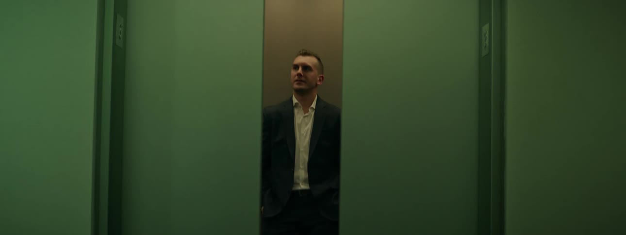 A man in a suit stands with his back against a wall in an elevator as the doors close