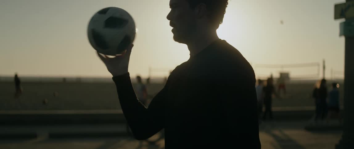 A man at the beach is spinning a soccer ball on his finger while people move behind him