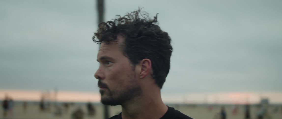 Man standing in park holds a soccer ball and looks around as people in background move along beach and road