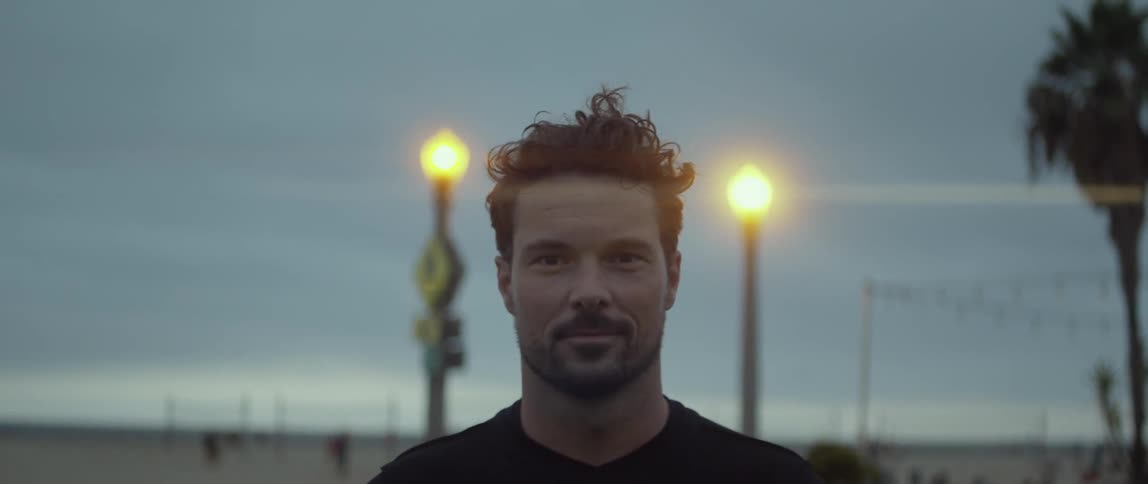 A man smiles as he holds a soccer ball while other people run and play volleyball on the beach behind him