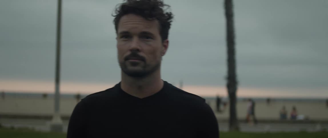 A man is standing outside as many people are walking and playing on the beach in the distance