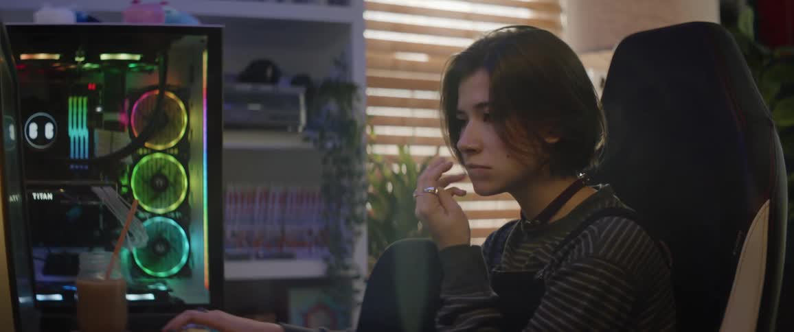 A teenage girl sits in front of a computer at home and bites her fingers and nods her head