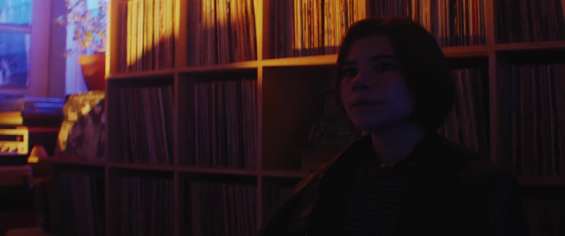 A woman standing in front of shelves holding record albums dances and sways and colorful lights flash outside the window