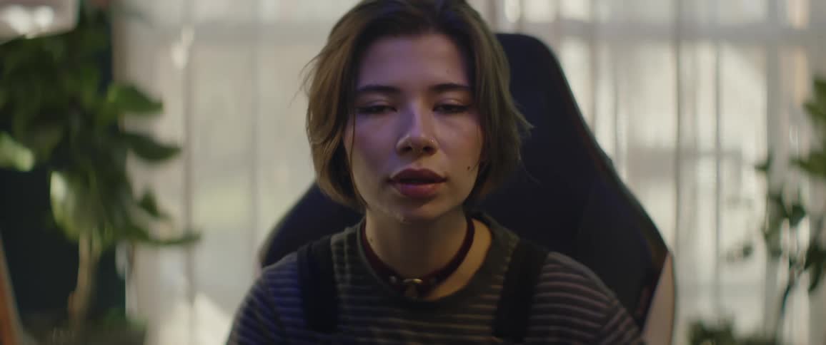 A young female sits in a chair in front of white curtains while she combs her fingers through her hair and looks into the camera