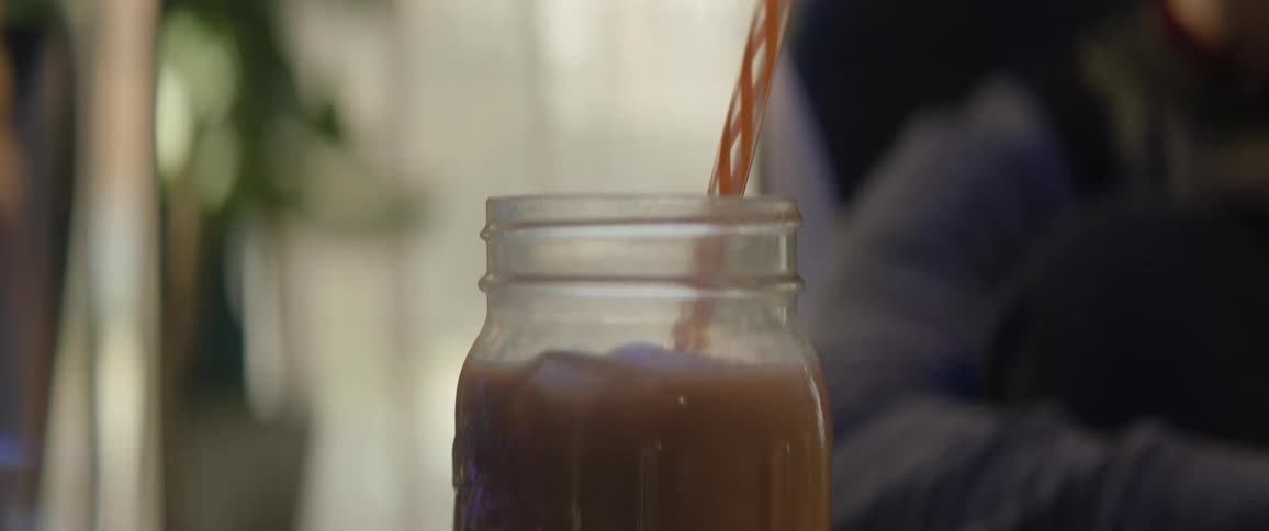 A girl sets a drink on a table and puts a straw in it and she sits down then picks it up and takes a drink