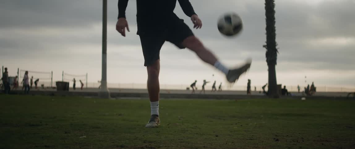 A man is outside in a field playing with a soccer ball and bouncing it off of his feet as people pass by on the street