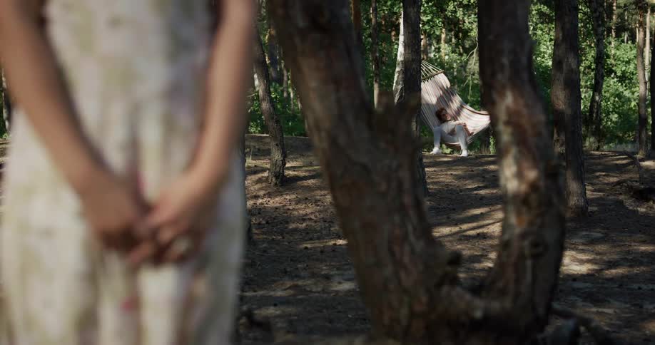 Woman stands near tree as man lies in swaying hammock in the background