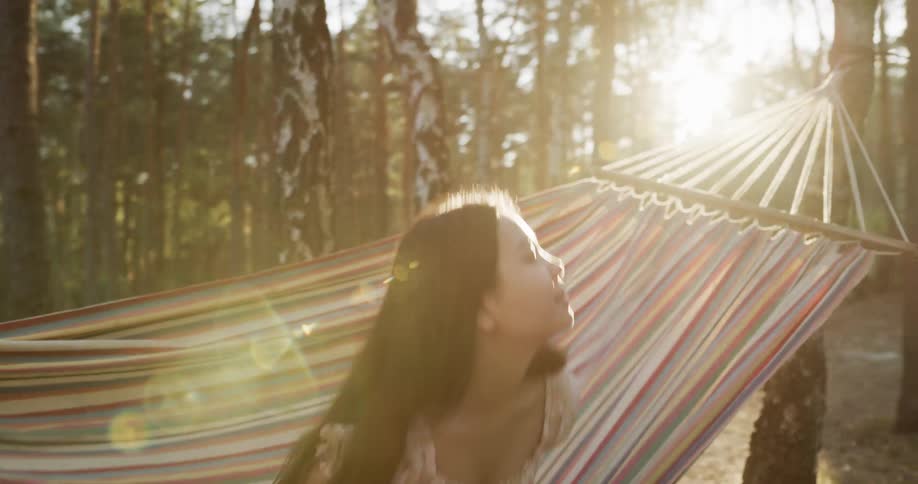 A woman wearing a dress in a forest walks to a hammock and sits down