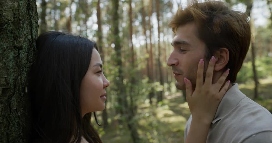 A woman leans against a tree as her boyfriend stands in front of her looking into her eyes and she puts her hand on his face and moves it over his heart