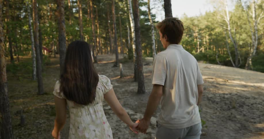 A man and a woman are holding hands as they walk into the woods as the sun shines through the trees