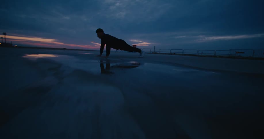 A man is outside near the beach doing push ups under a cloudy sky as the sun has set