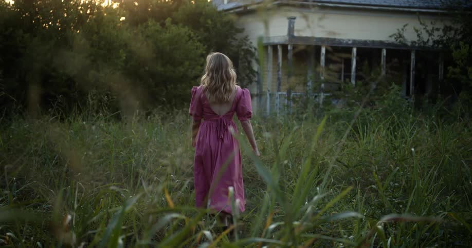 Woman walks through overgrown yard to old unmaintained house under a red sky