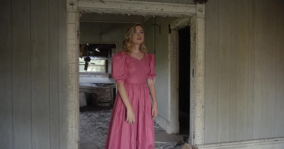 Blonde woman in a pink dress walks through a dilapidated building and reaches up towards the ceiling