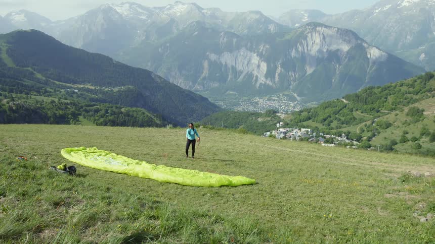 Woman stands with a paragliding canopy in high meadow overlooking town in valley between several mountains