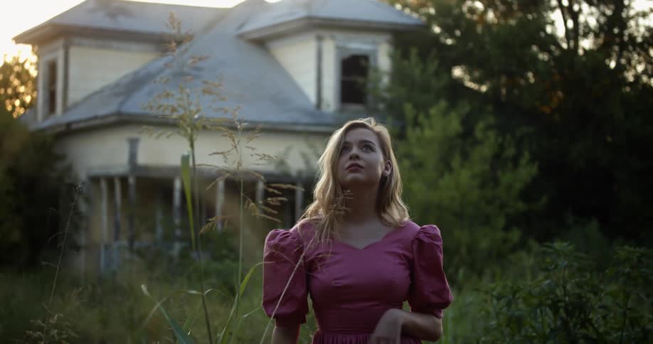 A woman poses behind weeds in front of a dilapidated home