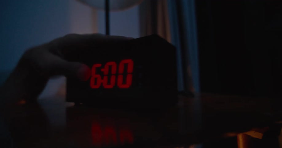 An alarm clock on the table is set at six oclock when someone in bed reaches over and turns it off