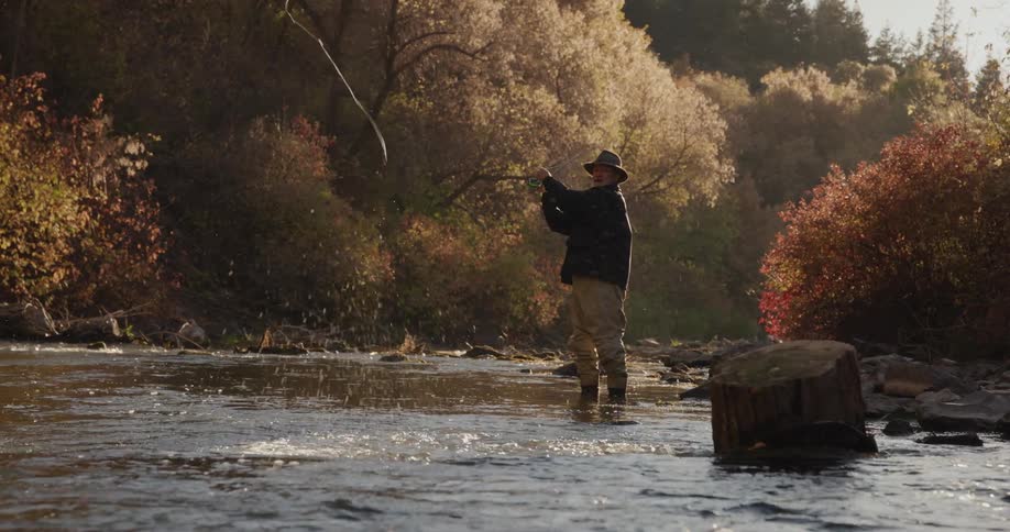 A man is standing in the woods along the side of the river as he casts his fishing line into the water