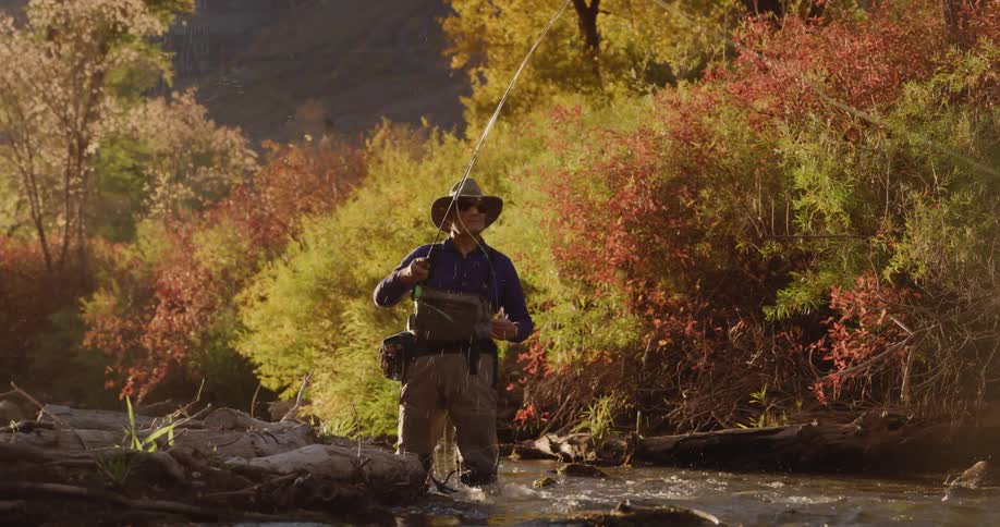 Elderly man with hat sunglasses and fishing net casts fishing line in the stream next to autumn forest
