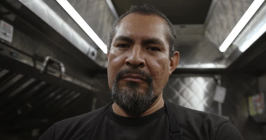 Male cook with goatee black shirt and apron stares at camera in the kitchen
