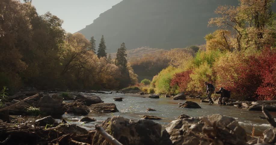Two friends wearing hats sunglasses jacket and pants sits on rock side by side fish in a stream near the forest with brown trees