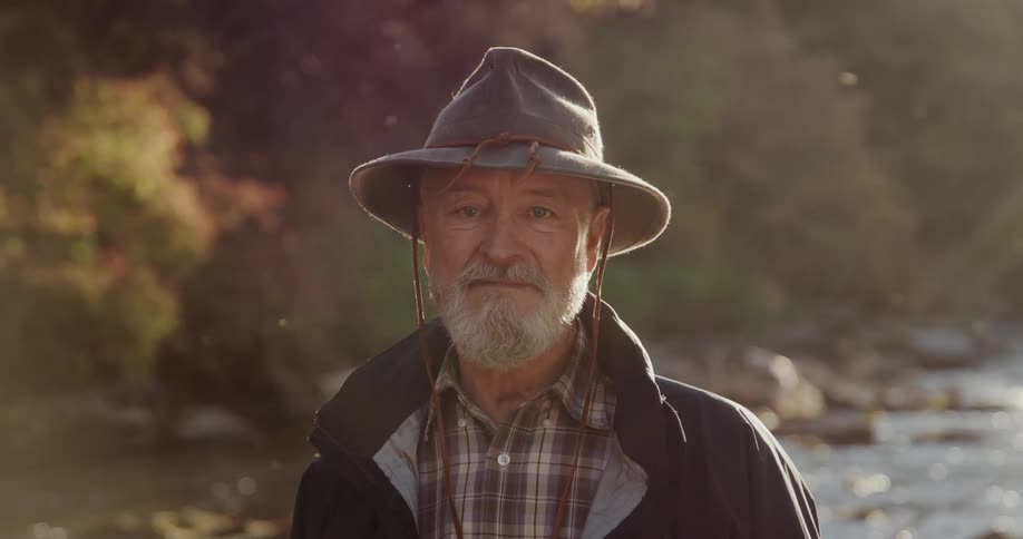 A graying fisherman looks into the camera while standing in front of a river during the day