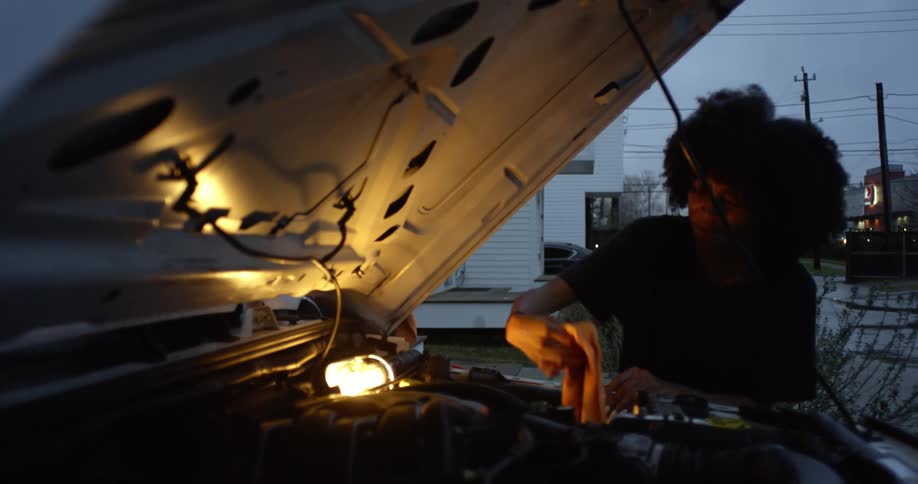 A woman is working under the hood of a truck as a man leans against the vehicle talking to her