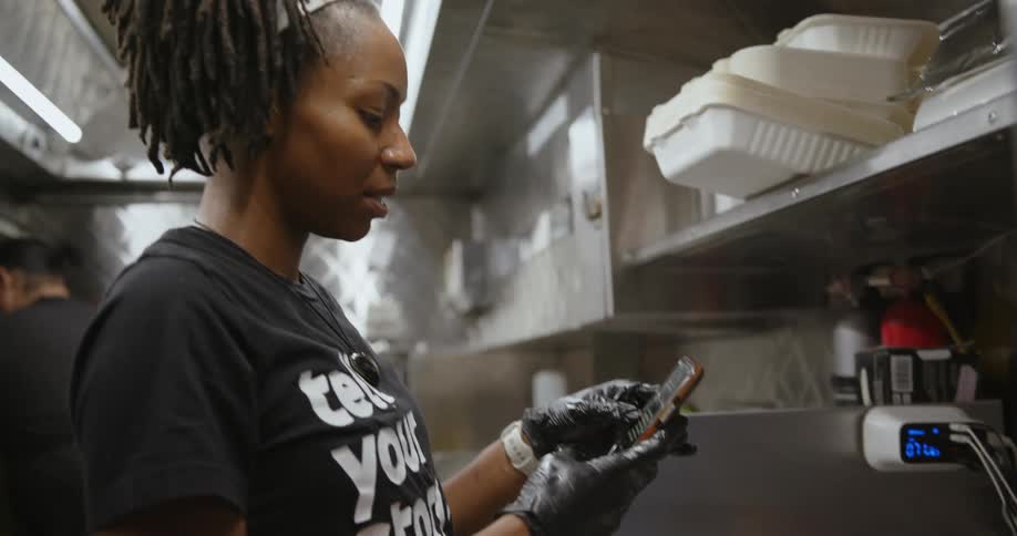 A woman working in a restaurant scrolls on her phone as a man behind her cooks