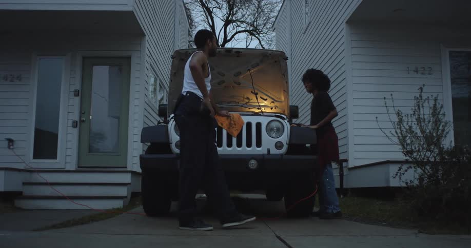 A man and a woman looking at the engine of a jeep parked at home in the morning