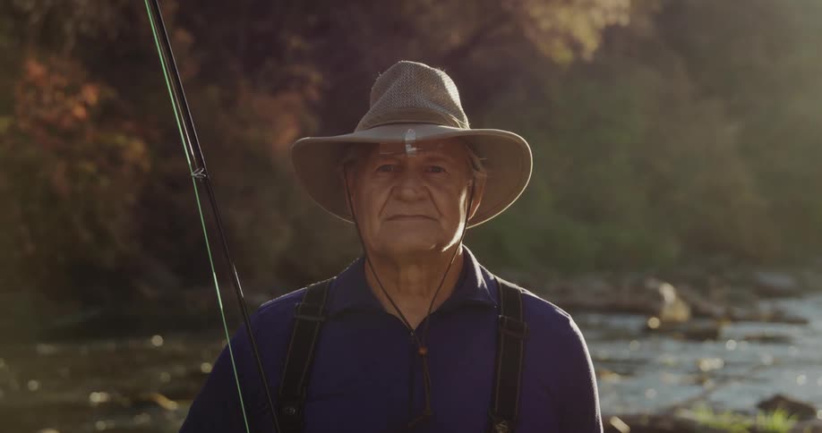 Elderly man wearing brown hat and blue shirt holds fishing rod in the forest stream