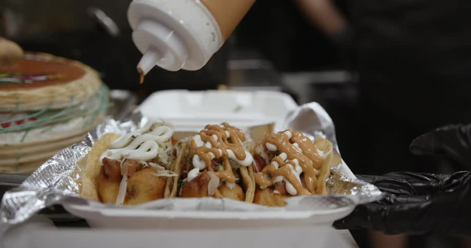 Three tacos are in a plastic container as someone is covering them with sauce