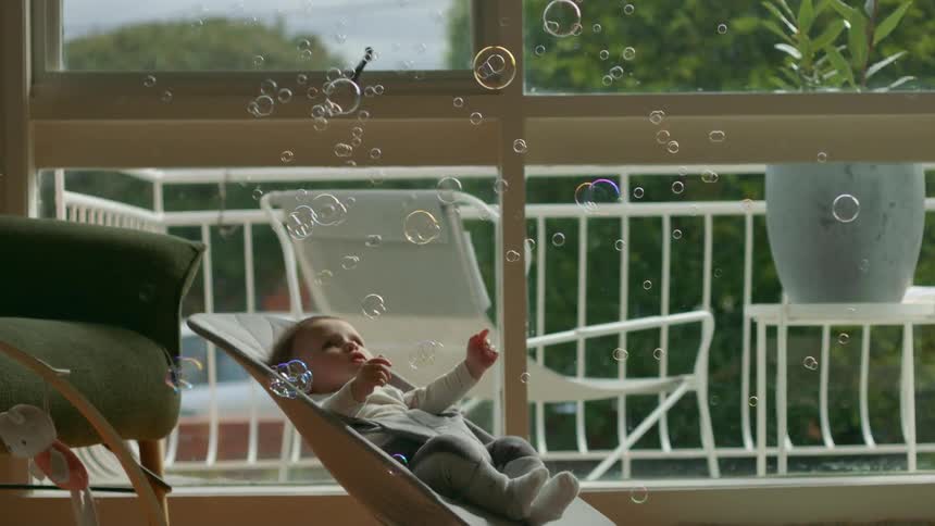An infant in a bouncing chair looks upwards as bubbles fall down around it