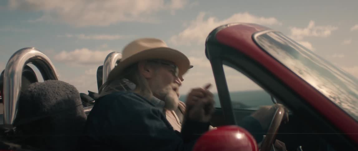 An elderly couple is driving along a coast as they smile and talk