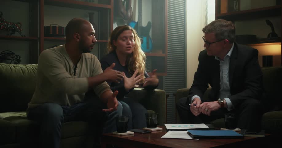 A biracial couple sits on a couch while having a discussion with a caucasian man that points to paperwork