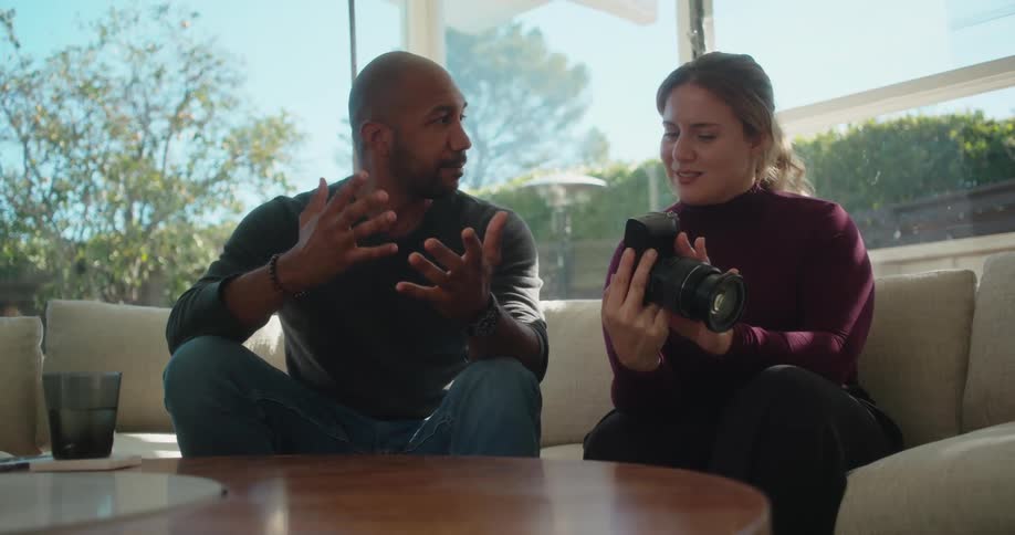 Woman with brown ponytail crimson sweater and black pants sits on beige couch next to bald man wearing gray sweatshirt and jeans checking camera in the living room at home