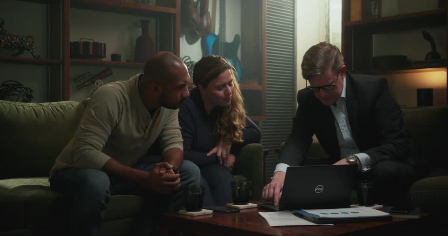 Three people are sitting in a living room talking and looking at a laptop computer on the table
