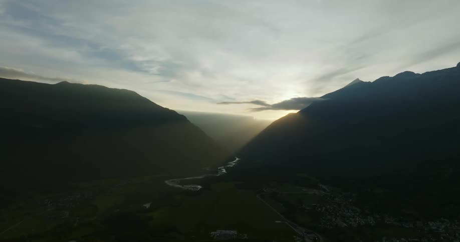 Sun shines from around mountain into shadowy valley containing river and town