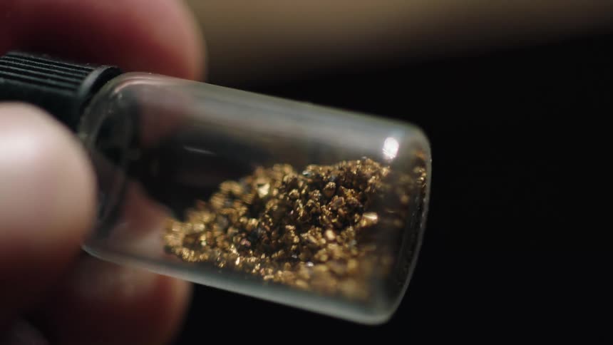 A man is holding a small jar containing gold granules in his fingers and he is turning it so they move