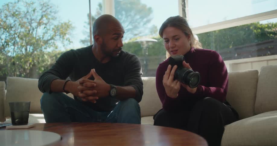 A man and a woman are sitting in the living room and she is holding a camera and showing him pictures as they talk