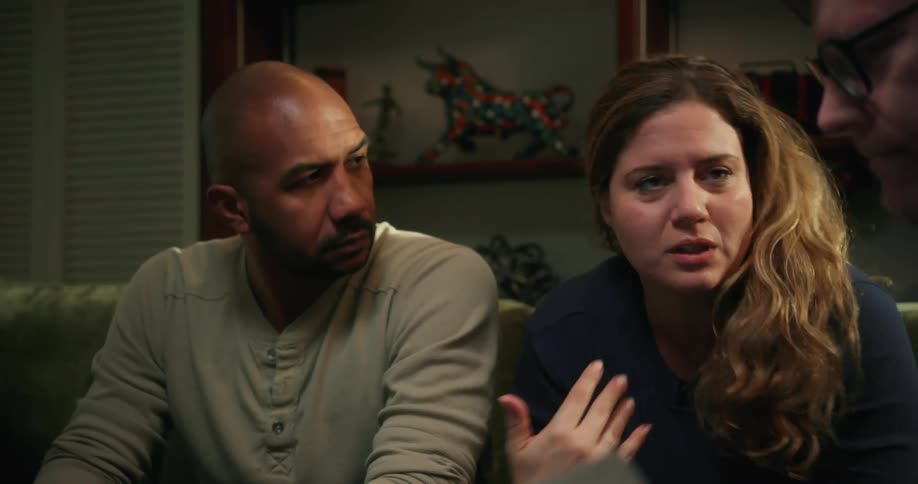 Three people are sitting in a living room talking and the woman is moving her hands as she speaks