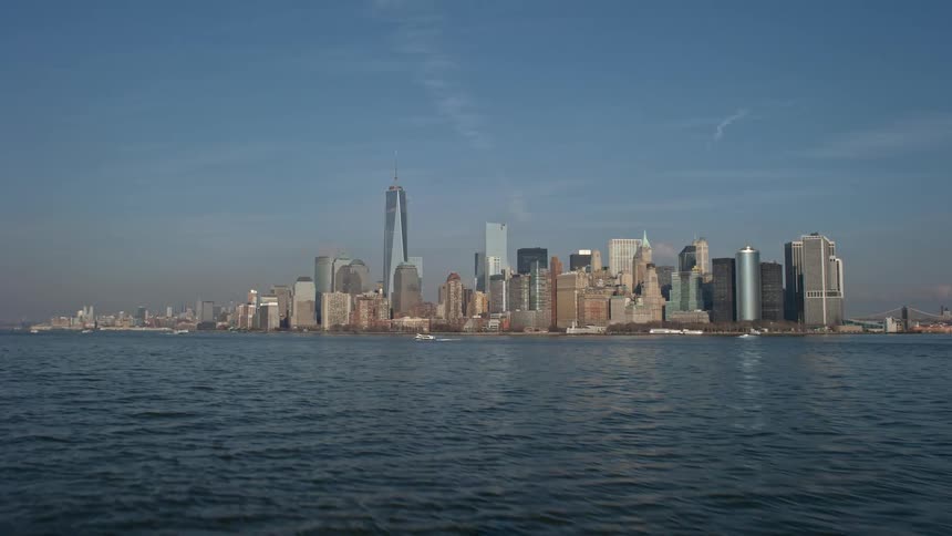 Approaching and revealing the Lower Manhattan skyline from New York Harbor with multiple tall buildings and skyscrapers towering over the city on a sunny winter day