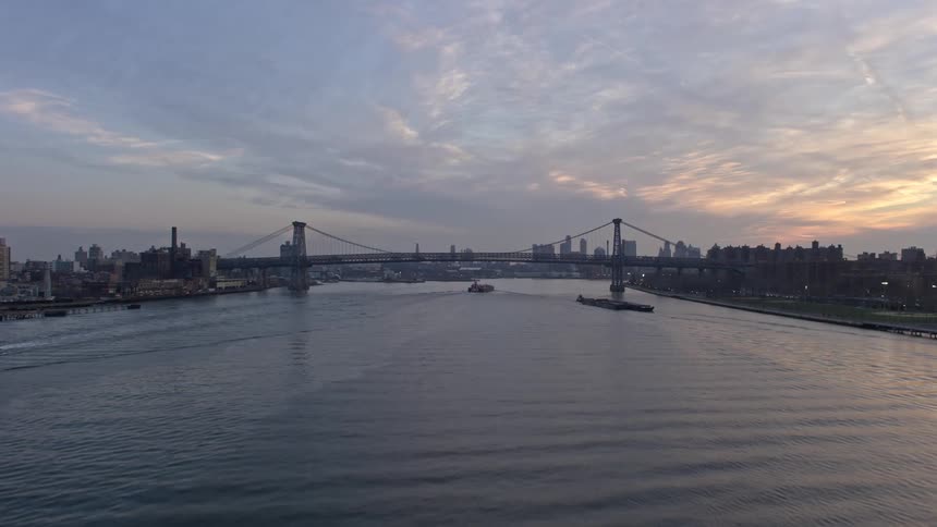 Approaching barges sail under Williamsburg Bridge during sunset from a 5K aerial view of the East River in winter