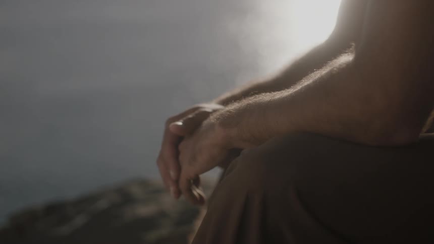 A shirtless man sits on a cliff near the ocean and contemplates things