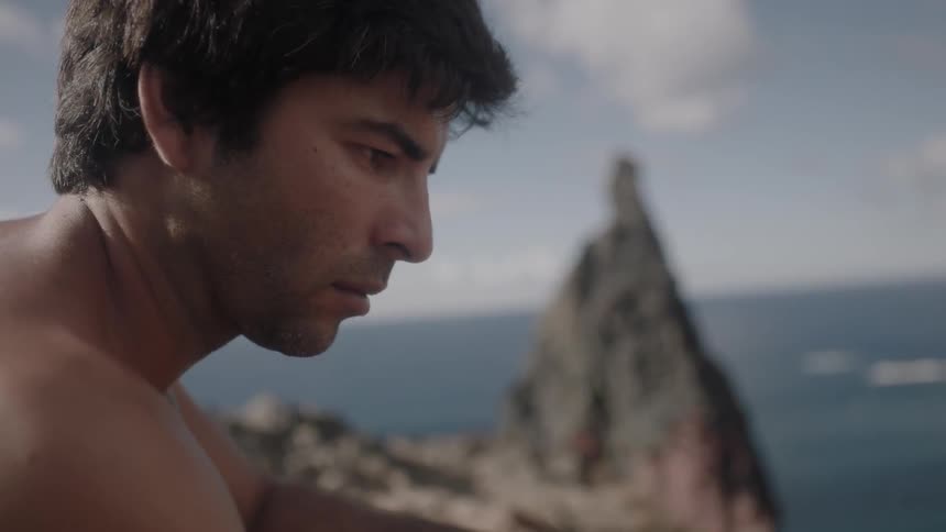 A shirtless man sits on a cliff near the ocean
