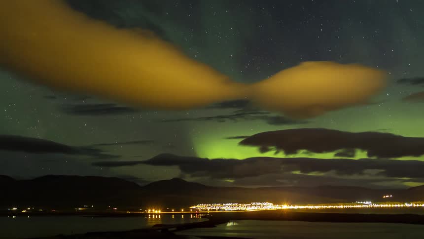 Vehicle lights streak across water and town under aurora borealis